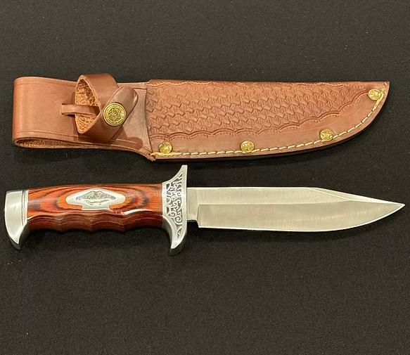 Bowie knife with engraved guard, wooden handle, and detailed leather sheath on black background.